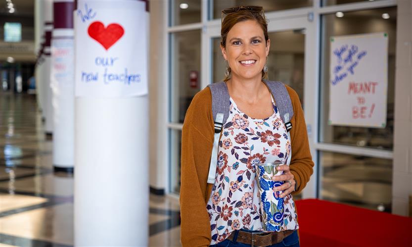 A woman wearing a floral shirt and brown cardigan smiles while standing in a school hallway
